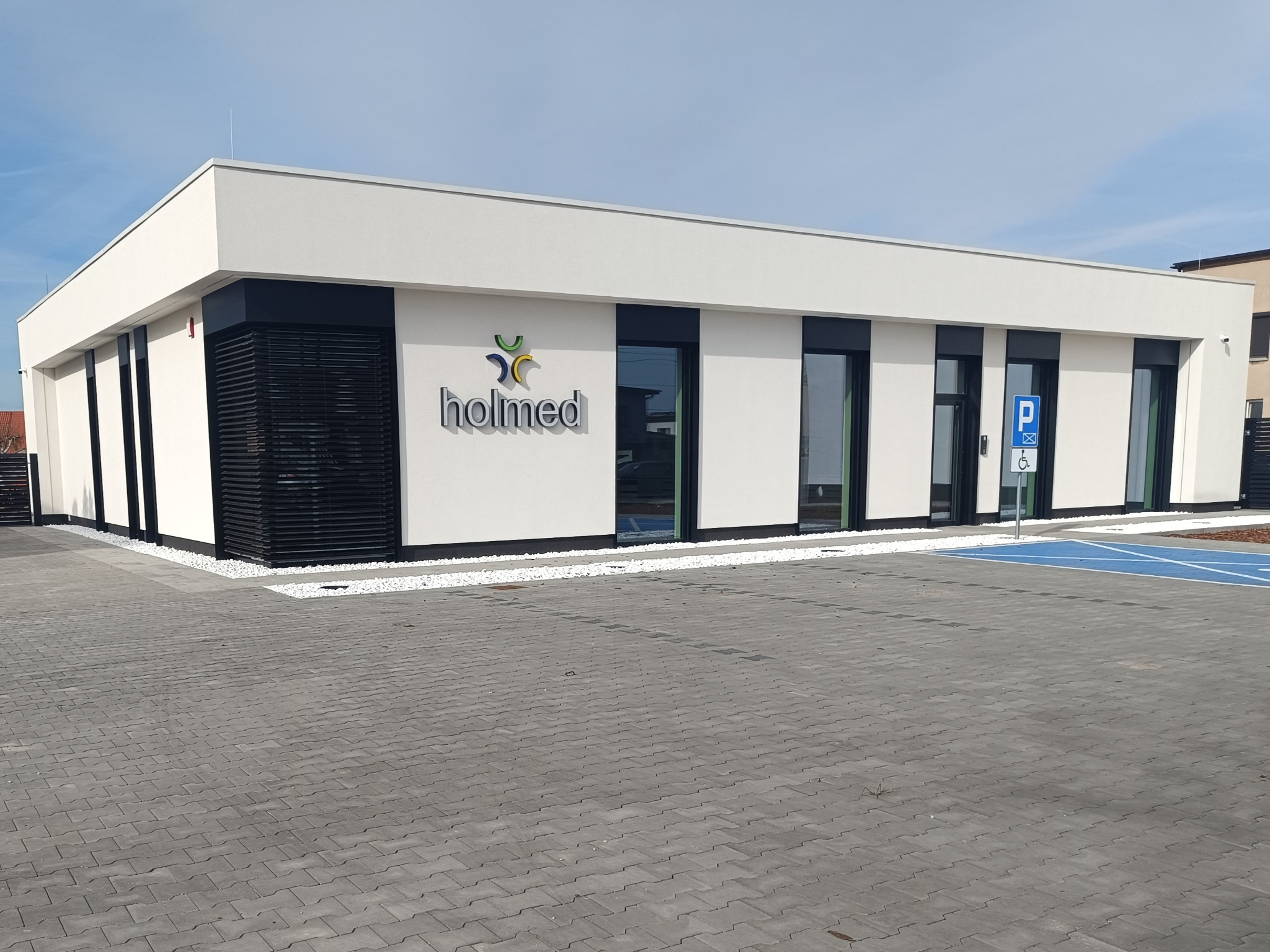 Nowoczesny budynek biurowy z dużymi oknami w czarnych ramach, z napisem 'holmed' nad wejściem, otoczony białymi kamykami i szarą kostką brukową na parkingu.