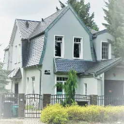 Szary dom jednorodzinny z dachem dwuspadowym i tabliczką adresową '17 Muszyńska', otoczony zielenią i ogrodzeniem z metalowymi przęsłami.