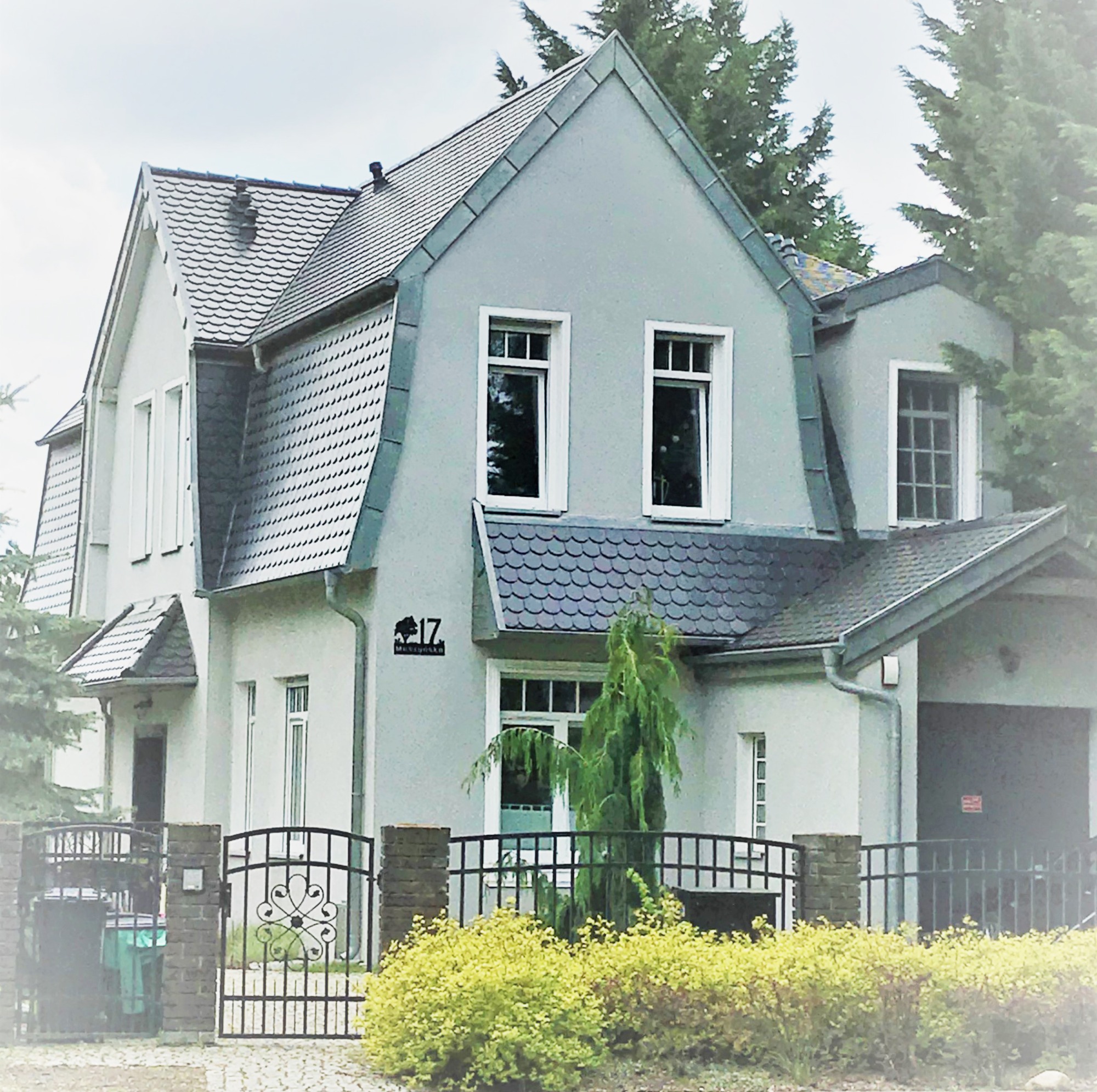 Szary dom jednorodzinny z dachem dwuspadowym i tabliczką adresową '17 Muszyńska', otoczony zielenią i ogrodzeniem z metalowymi przęsłami.