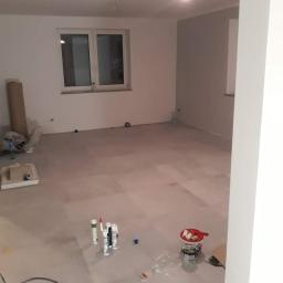 Marbud - Pokój w trakcie prac wykończeniowych: szpachlowane ściany, przygotowane materiały budowlane, widoczne okno i surowe podłoże pod podłogę.