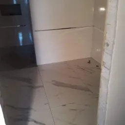 Świeżo wykafelkowana łazienka z białymi płytkami imitującymi marmur na podłodze i ścianach kabiny prysznicowej, widoczne srebrne listwy dekoracyjne i surowy narożnik ściany przed wykończeniem.