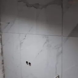 Ściana w łazience wyłożona białymi płytkami imitującymi marmur, widoczne otwory na gniazdka elektryczne i rury instalacyjne, etap prac wykończeniowych.