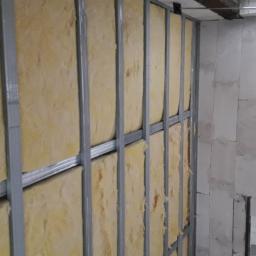 Marbud - Montaż izolacji z wełny mineralnej w metalowym stelażu na ścianie z betonu komórkowego, widoczne elementy instalacji pod sufitem.