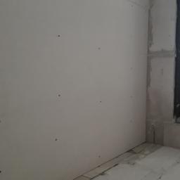 Marbud - Ściana wewnętrzna z zamontowaną płytą gipsowo-kartonową, widoczne wkręty mocujące, surowy stan wykończenia przy oknie, fragment posadzki w trakcie prac.