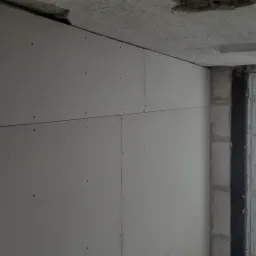 Ściana z zamontowaną płytą kartonowo-gipsową, widoczne śruby mocujące, surowy sufit i okno z widokiem na zewnątrz.