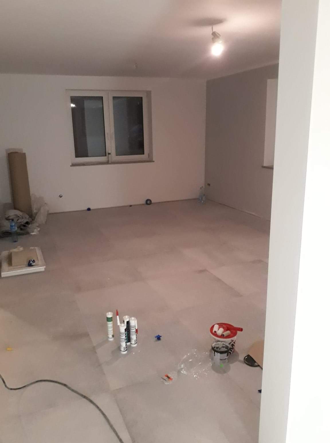 Pokój w trakcie prac wykończeniowych: szpachlowane ściany, przygotowane materiały budowlane, widoczne okno i surowe podłoże pod podłogę.
