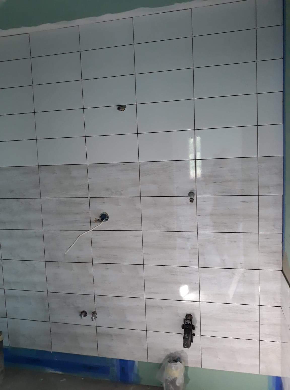 Ściana w trakcie układania płytek, widoczne otwory na instalacje, dolny rząd płytek o wzorze imitującym drewno, zabezpieczona podłoga.