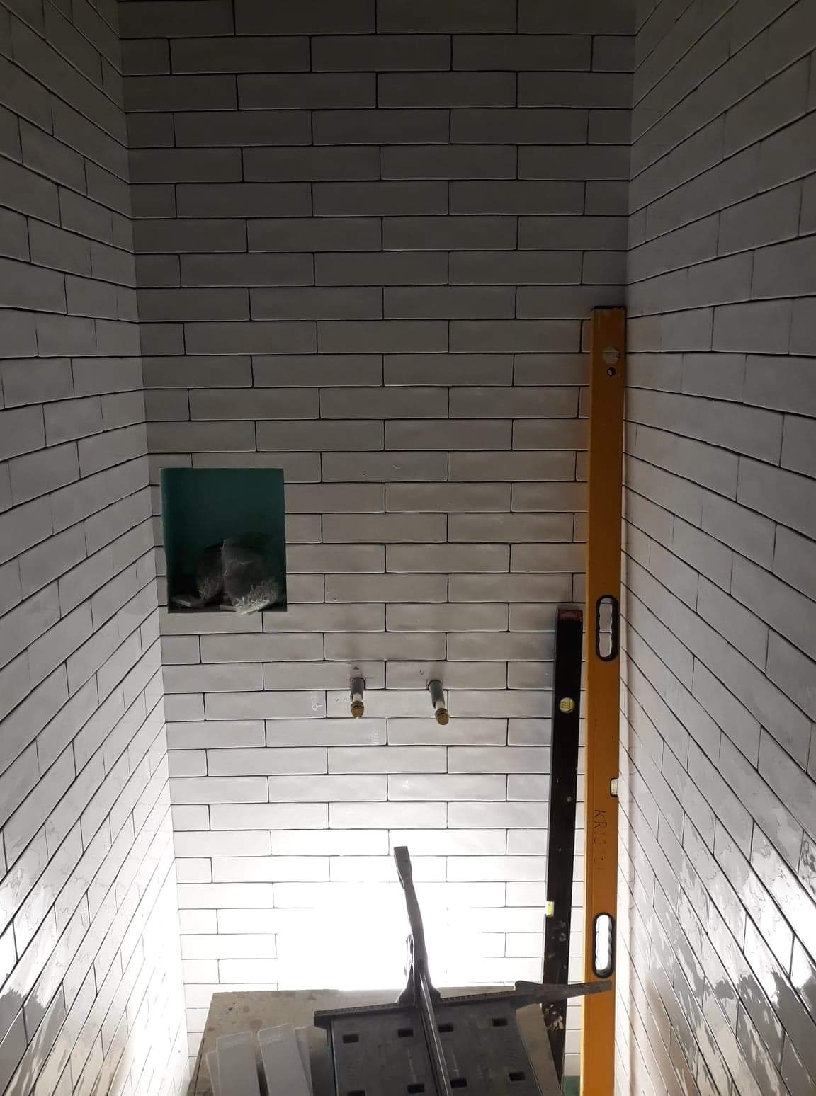 Świeżo wyłożona, jasnoszara, prostokątna glazura w małej łazience, z wnęką na kosmetyki, widoczne wystające rurki instalacji wodnej, poziomica przy ścianie i narzędzia glazurnicze na podłodze.