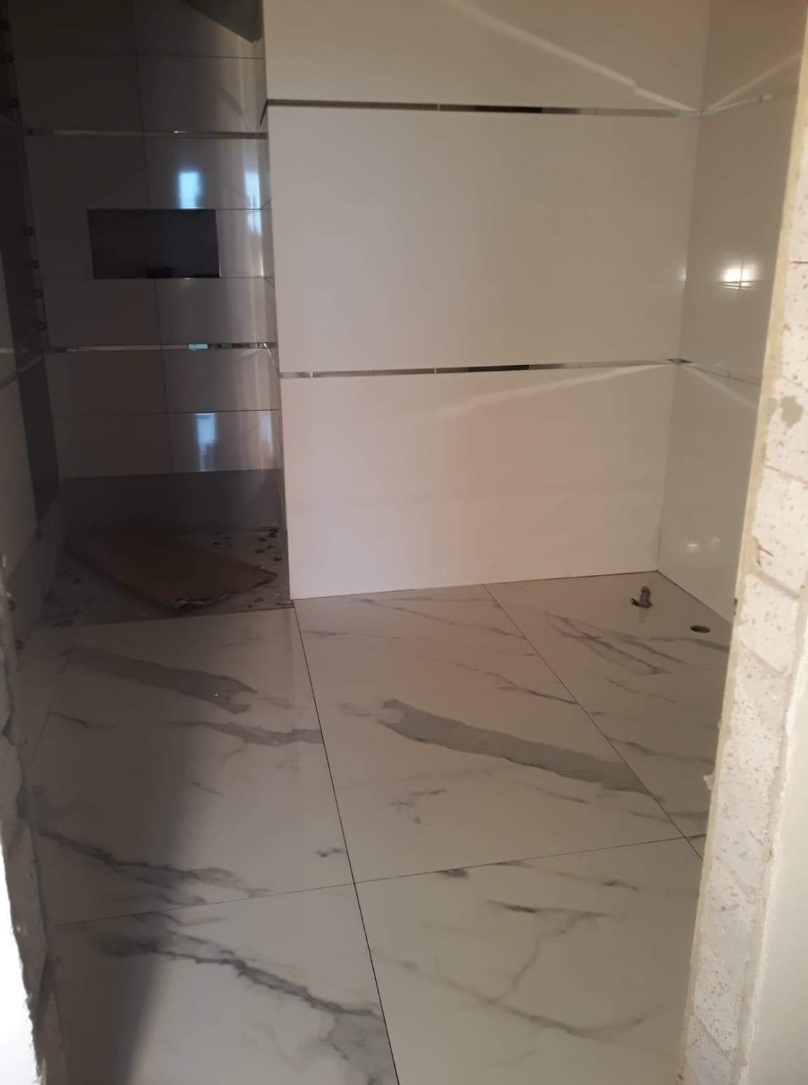 Wnętrze łazienki w trakcie prac wykończeniowych, widoczne białe płytki marmurowe na podłodze i ścianach z dekoracyjnymi, metalowymi listwami. Po lewej stronie wnęka z ciemniejszymi płytkami.