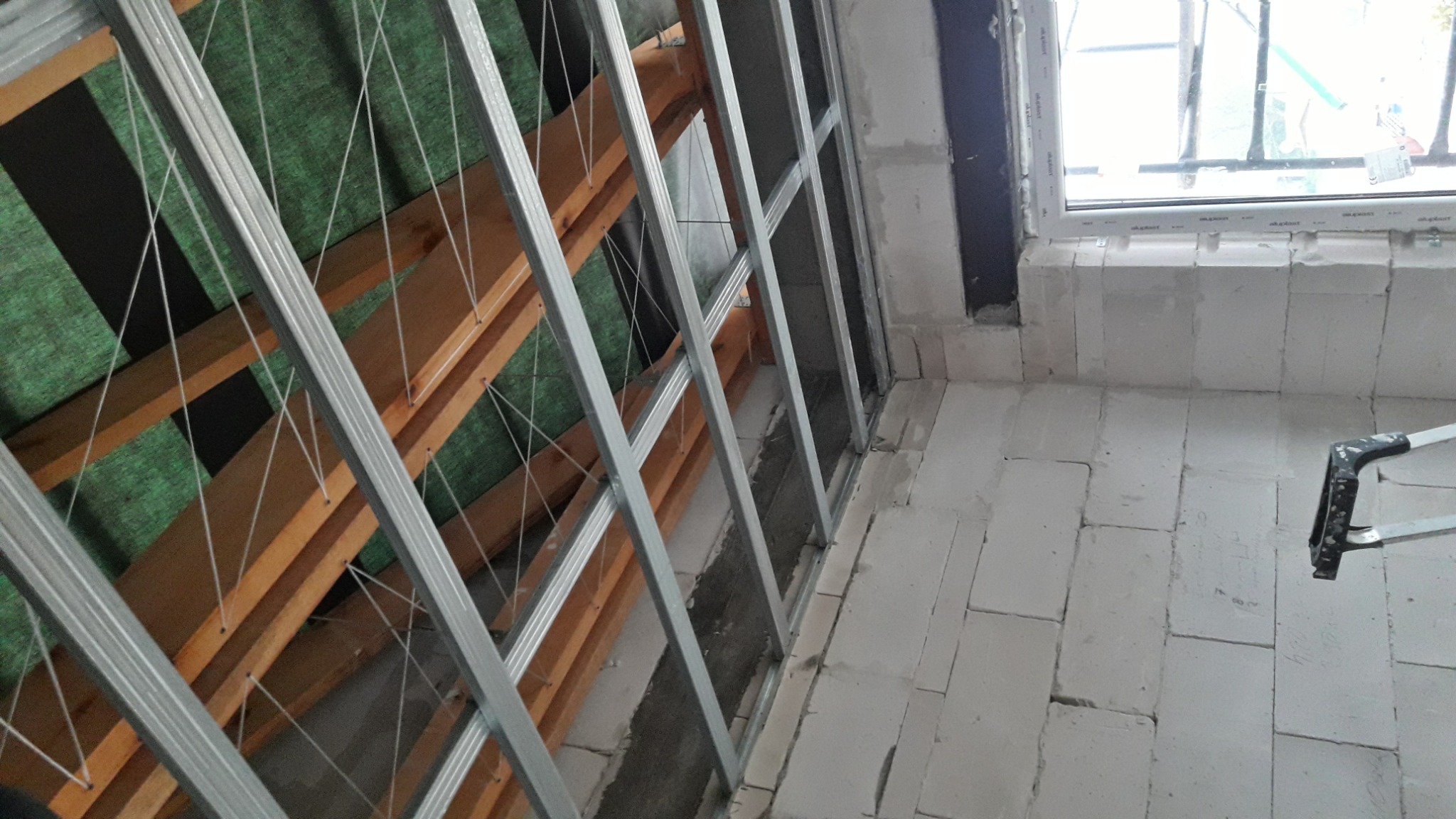 Konstrukcja szkieletowa z metalowych profili i drewnianych łat na zielonej macie, przygotowana pod zabudowę płytami karton-gips w pomieszczeniu z oknem i surowymi ścianami z białych bloczków.