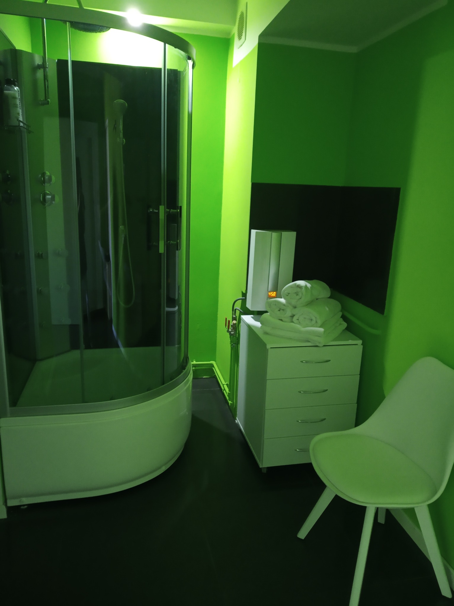 Nowoczesna kabina prysznicowa z hydromasażem, biała komoda z ręcznikami i krzesło w gabinecie z zielonymi ścianami.