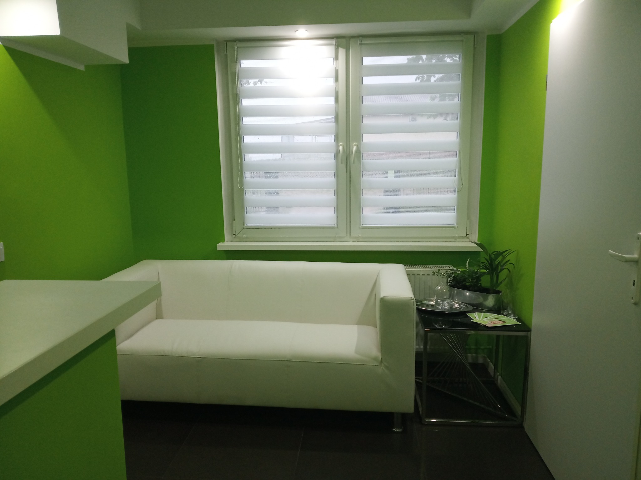 Poczekalnia w gabinecie z zielonymi ścianami, białą sofą, stolikiem ze szklanym blatem i rośliną doniczkową, okno z żaluzjami.