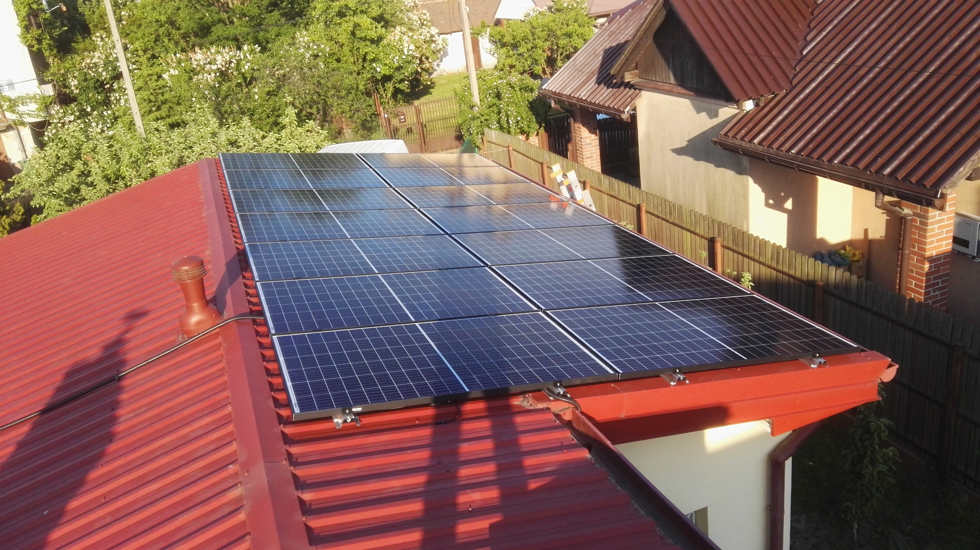 Panel słoneczny na czerwonym dachu z blachy trapezowej, widok z góry, cień przewodu na dachu, drzewa w tle.