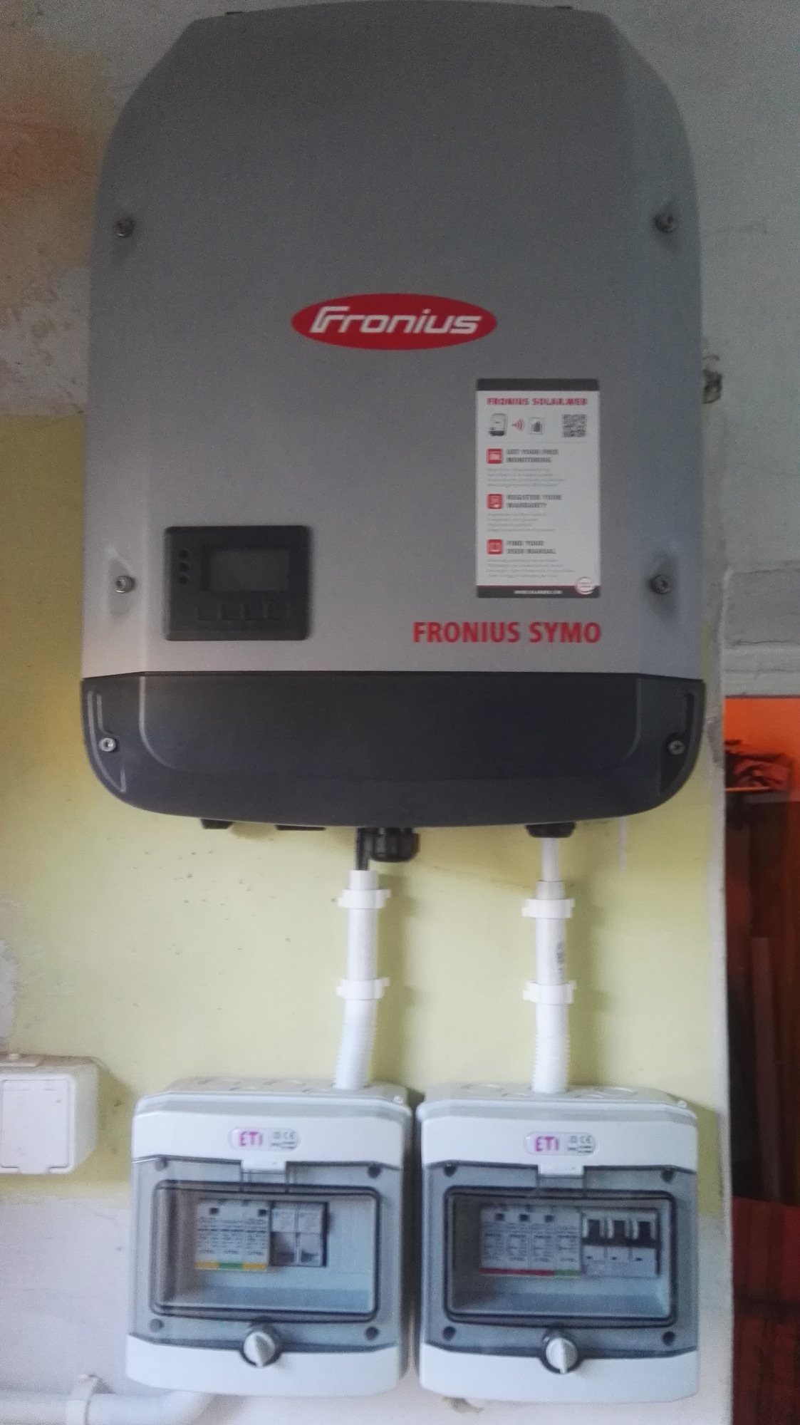 Zamontowany inwerter Fronius Symo z dwoma skrzynkami bezpiecznikowymi ETI na żółtej ścianie, instalacja wewnętrzna.