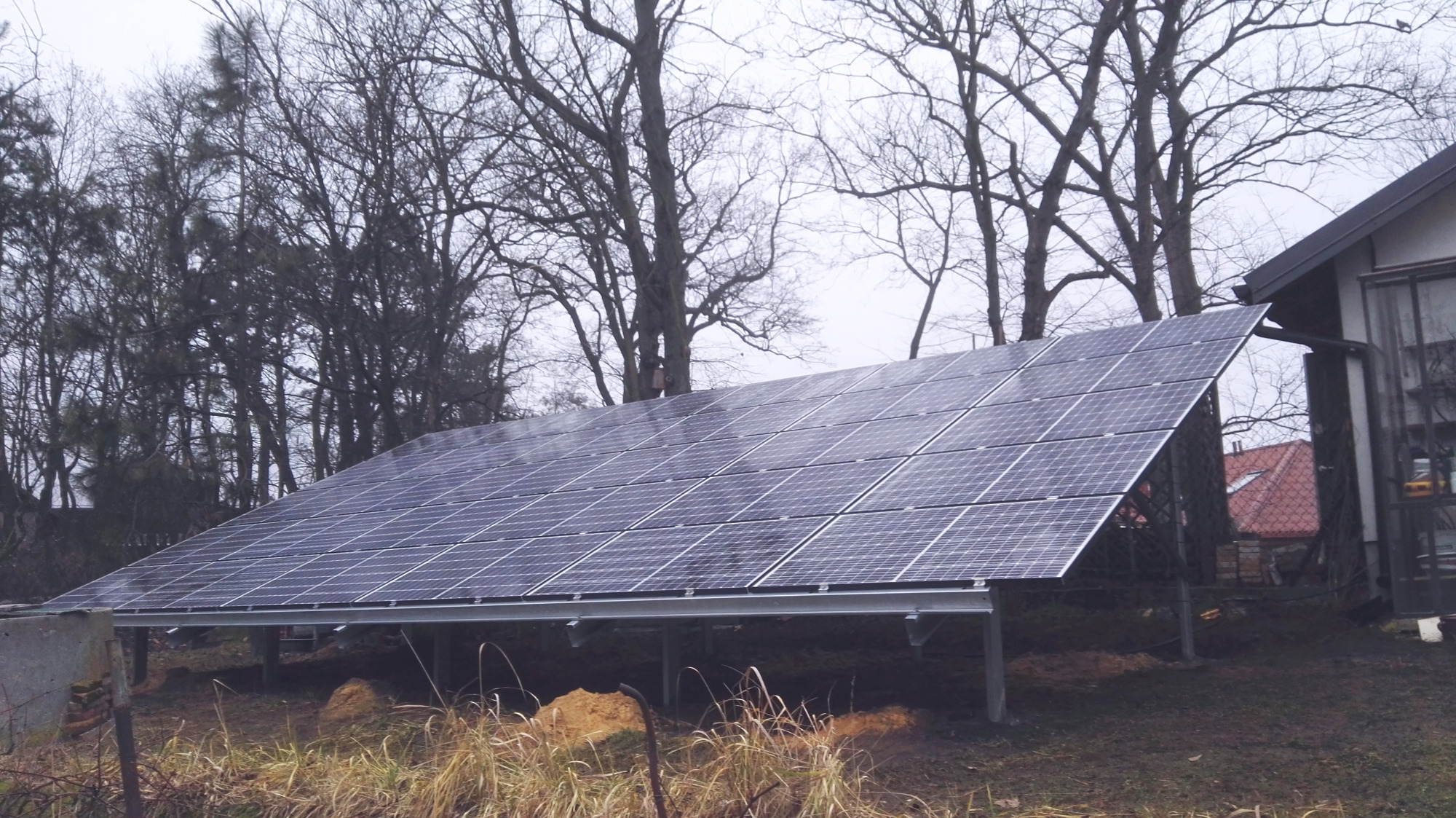 Instalacja paneli słonecznych na metalowej konstrukcji, widoczna z boku, na tle domu i drzew bez liści w pochmurny dzień.