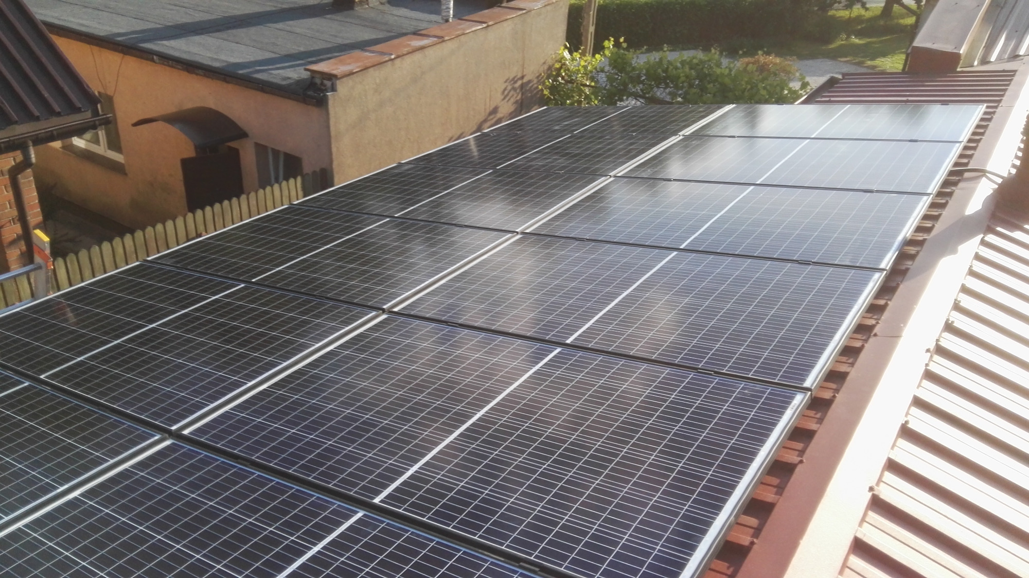 Panele słoneczne na dachu domu jednorodzinnego, widok z góry, w tle budynek z cegły i drewniany płot.