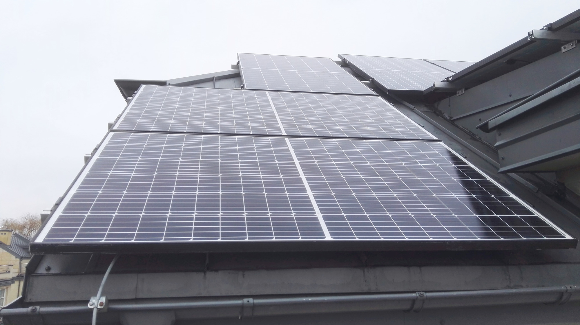 Panele fotowoltaiczne zamontowane na metalowym dachu budynku, widoczne przewody i elementy mocujące.