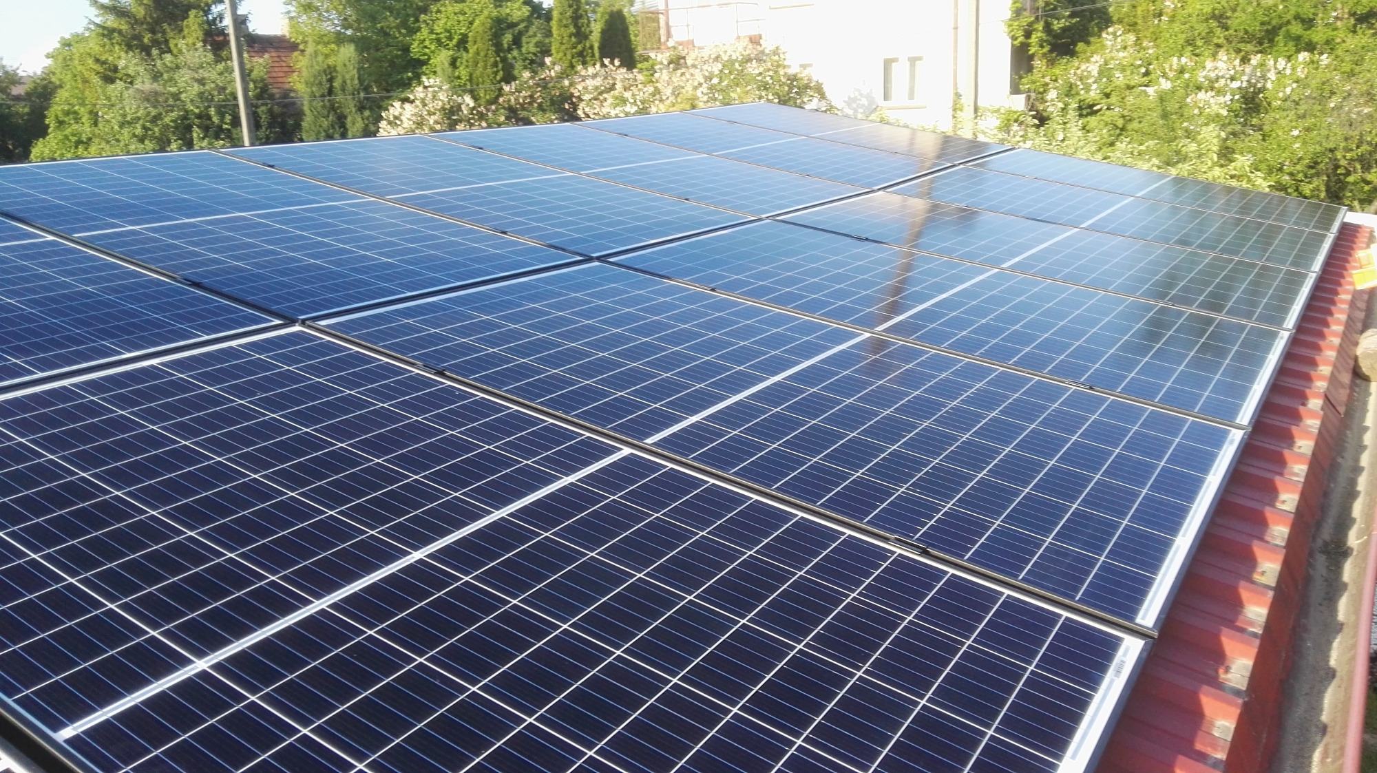 Panel słoneczny na czerwonym dachu, widok z góry, częściowo zacieniony, w tle drzewa i budynek.