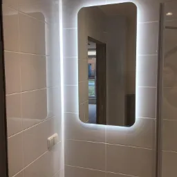 Nowoczesna łazienka z podświetlanym lustrem, białe płytki ceramiczne ułożone poziomo, widoczne baterie umywalkowe i włącznik światła.