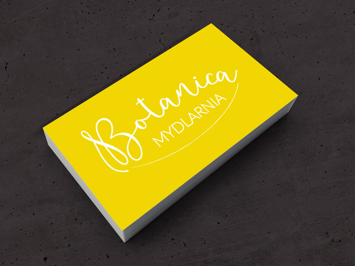 Żółty stos wizytówek z białym logo 'Botanica MYDLARNIA' na tle ciemnoszarej teksturowanej powierzchni.