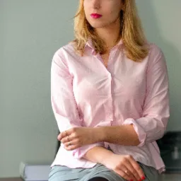 Portret kobiety w różowej koszuli i szarej spódnicy siedzącej na biurku, z zamyślonym wyrazem twarzy i spojrzeniem w bok.