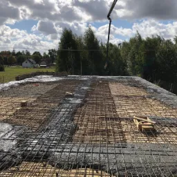 Wylewanie betonu na przygotowane zbrojenie i deskowanie stropu, widoczna pompa do betonu oraz otoczenie budynku z drzewami i zabudowaniami w tle.