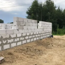 Wznoszenie muru z bloczków betonowych na zaprawie cementowej, widoczne wiązania, grunt piaszczysty, w tle samochód osobowy i zalesiony teren.