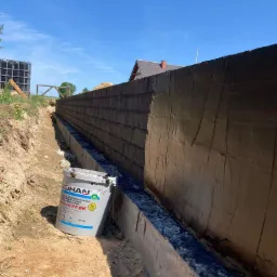 Świeżo zaizolowany fundament z widocznym wiadrem z masą hydroizolacyjną firmy Zohan, obok wykopu z ziemią i fragmentem krajobrazu z domem w tle.