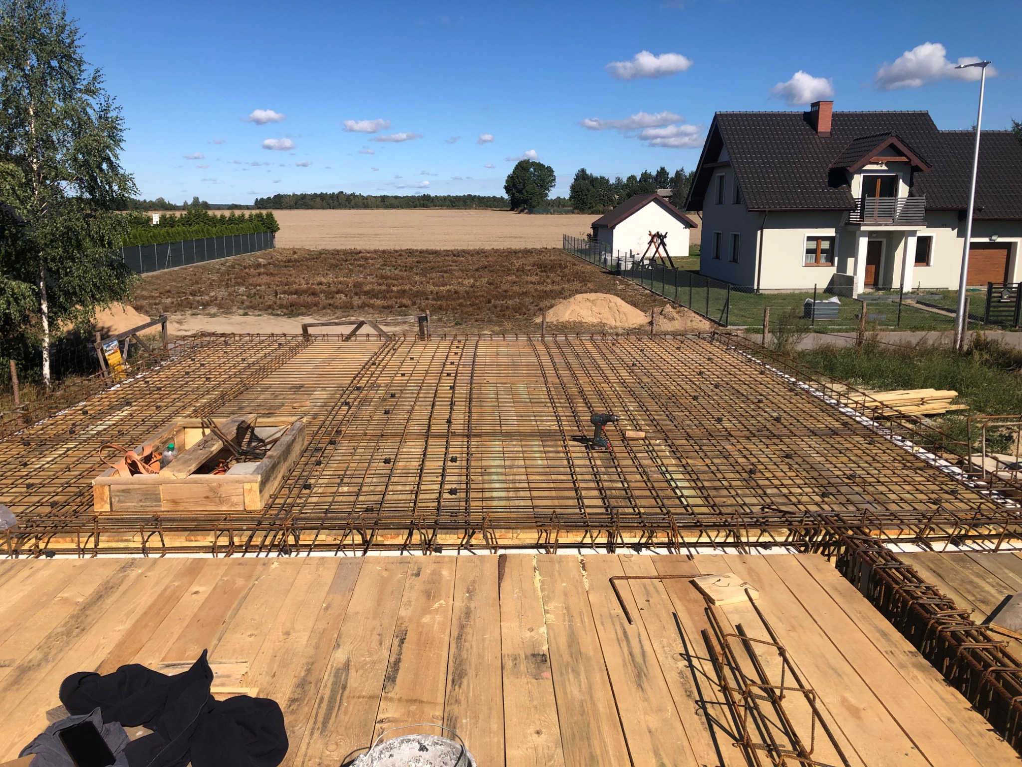 Zbrojenie płyty fundamentowej domu jednorodzinnego z widocznymi drewnianymi deskami szalunkowymi, stalowymi prętami zbrojeniowymi tworzącymi siatkę oraz drewnianą skrzynką na narzędzia; w tle dom...