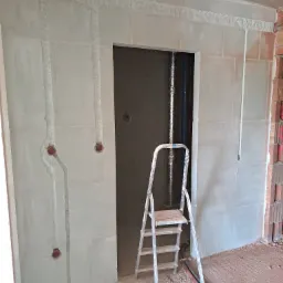 Surowy stan ścian z wyfrezowanymi bruzdami pod przewody elektryczne, widoczne puszki instalacyjne, drabina aluminiowa, kable na podłodze.