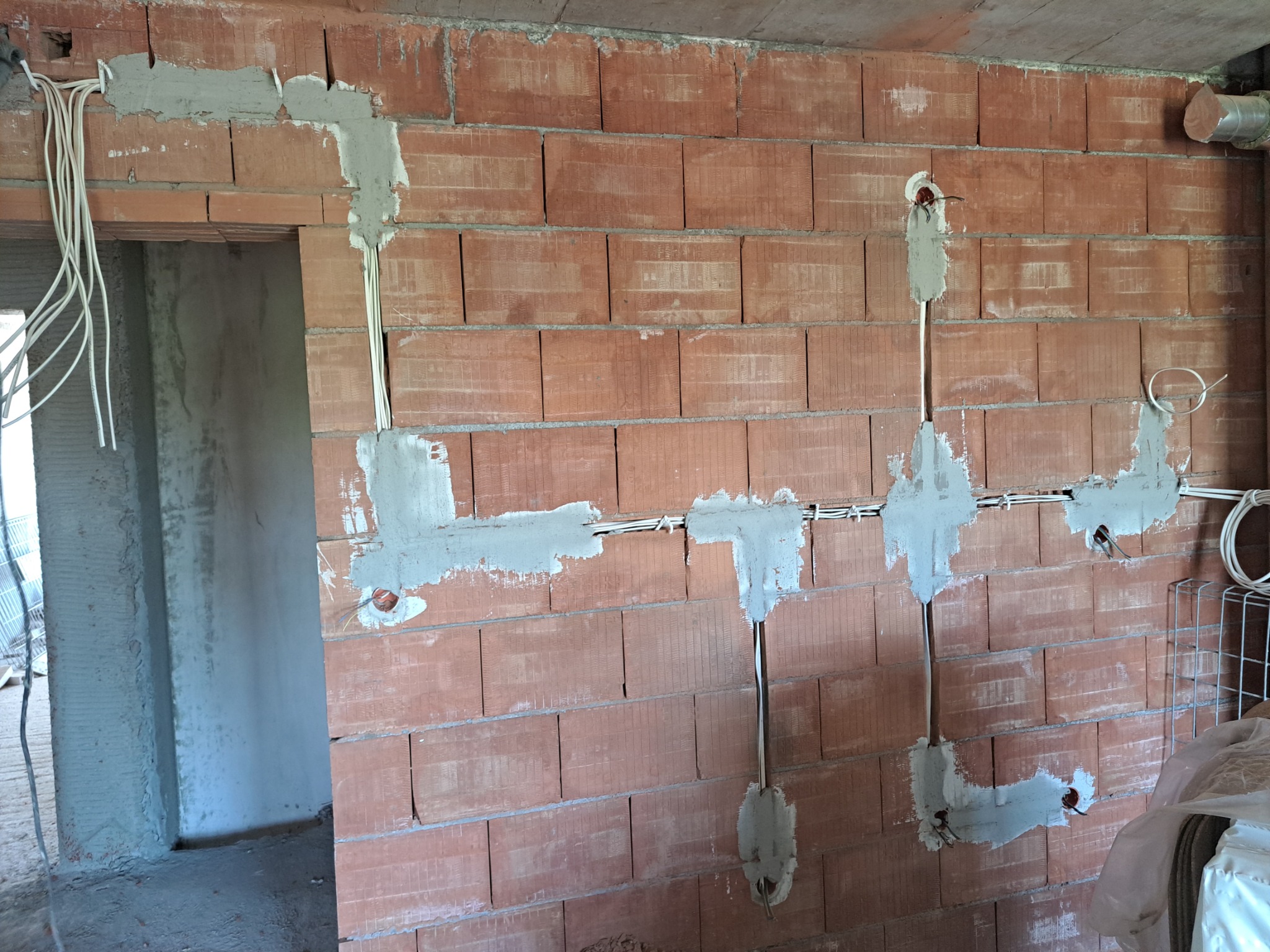 Surowa ściana z cegły z wyżłobieniami wypełnionymi gipsem, w których poprowadzone są białe przewody elektryczne. Widoczne puszki instalacyjne i wiszące kable przy otworze drzwiowym.