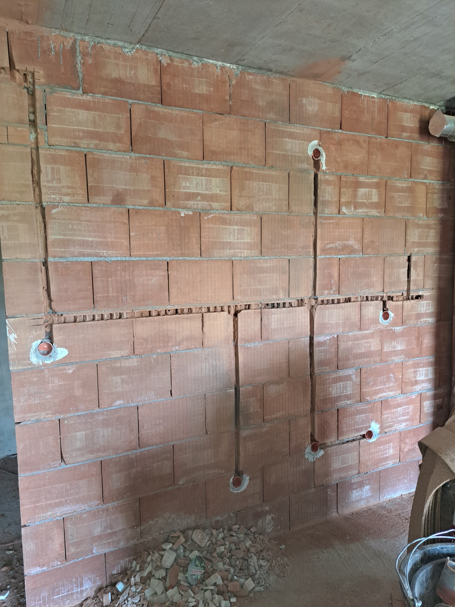 Murowana ściana z wyżłobionymi bruzdami i zamontowanymi puszkami instalacyjnymi pod instalację elektryczną, widoczne fragmenty gruzu na podłodze.