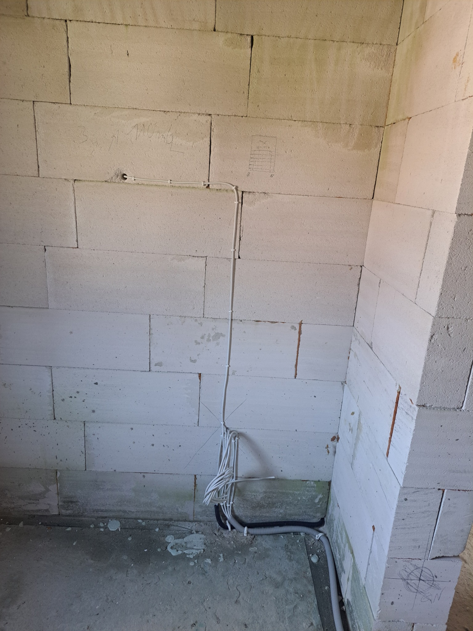Wewnętrzny róg surowego pomieszczenia z białych bloczków, widoczne kable elektryczne prowadzone po ścianie i w peszlu przy podłodze, surowy beton na podłodze.