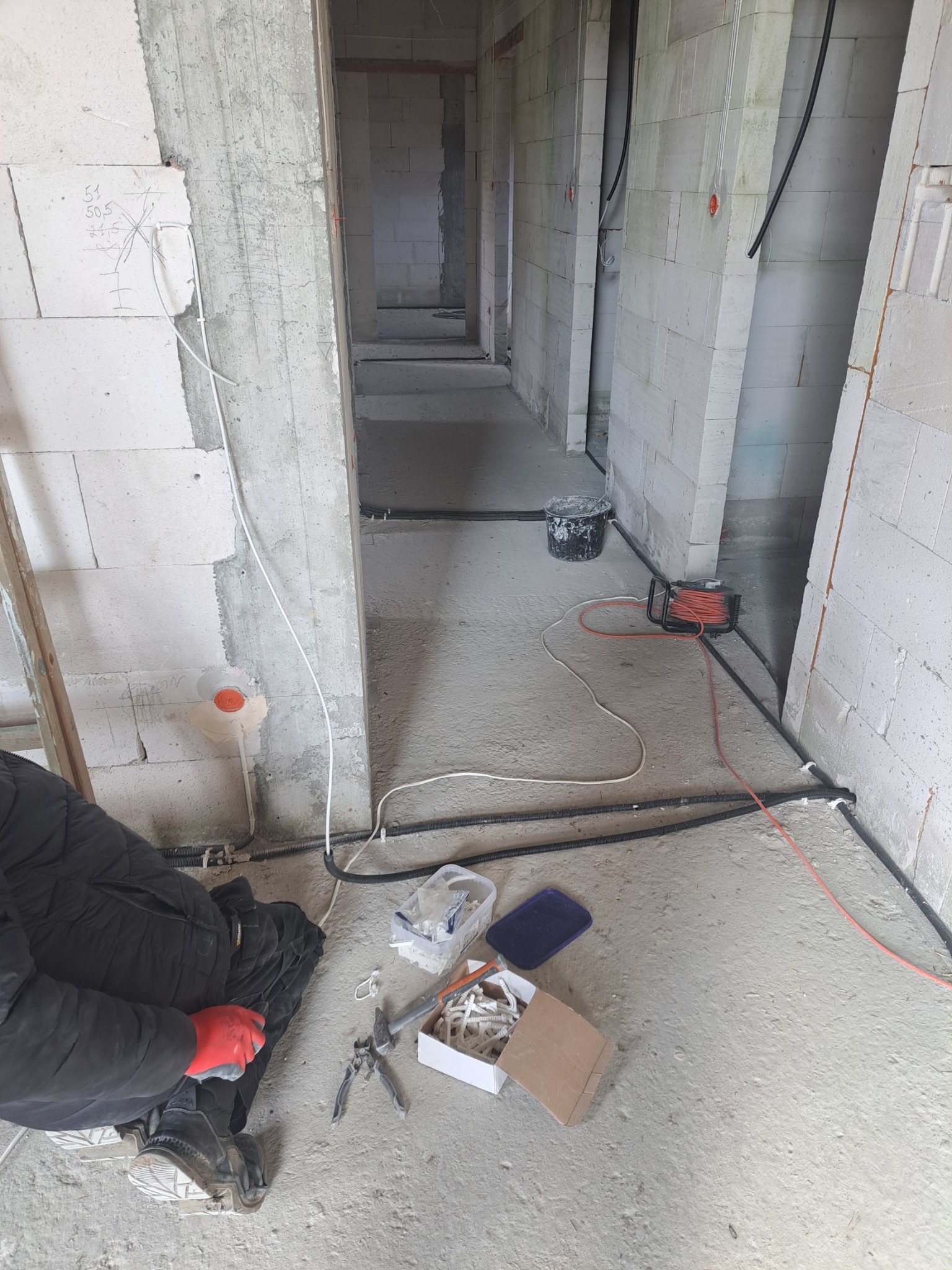 Elektryk w czerwonych rękawicach klęczy przy podłodze z narzędziami i puszkami, montując kable w surowym wnętrzu budynku z betonu komórkowego, widoczne oznaczenia na ścianie.