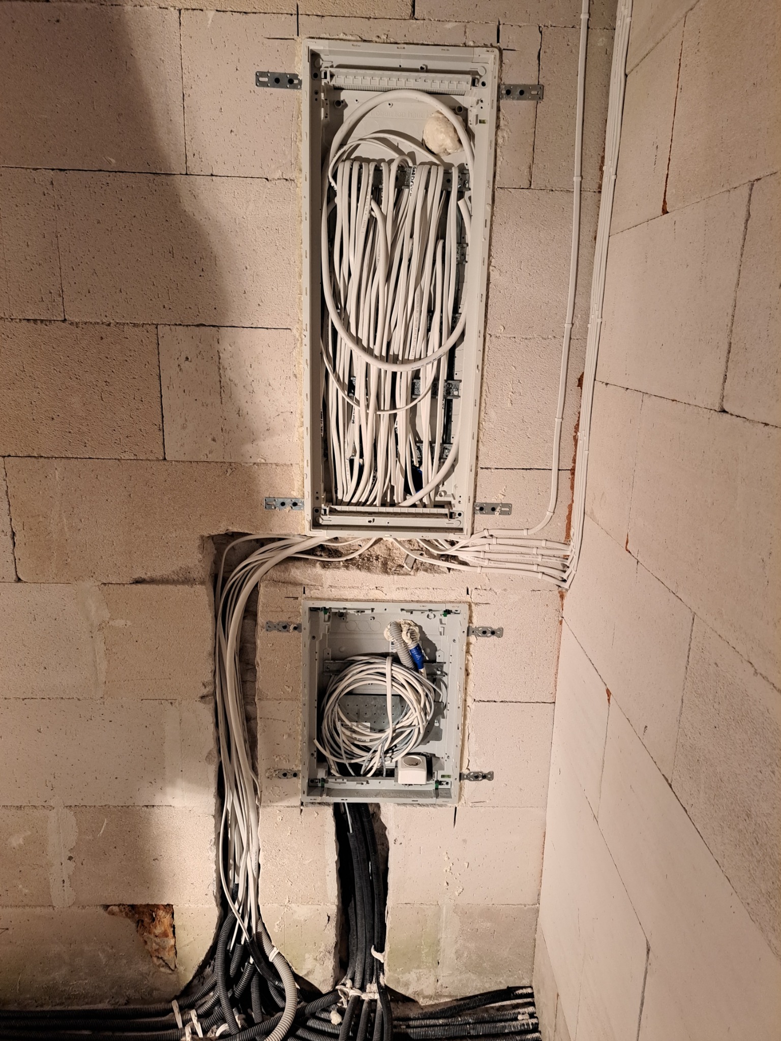 Dwa panele elektryczne w surowej ścianie z betonu komórkowego, górny panel wypełniony białymi kablami, dolny z białymi i niebieskimi, poniżej widoczne czarne rury karbowane.