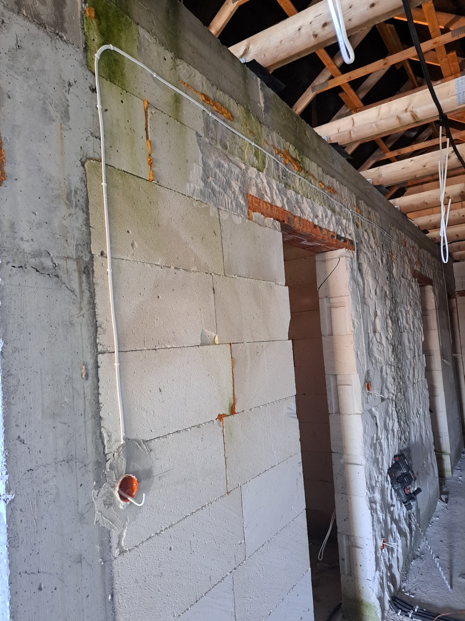 Montaż instalacji elektrycznej w surowym stanie: białe kable prowadzone po ścianie z betonu komórkowego, widoczna puszka elektryczna i fragment drewnianej konstrukcji dachu.