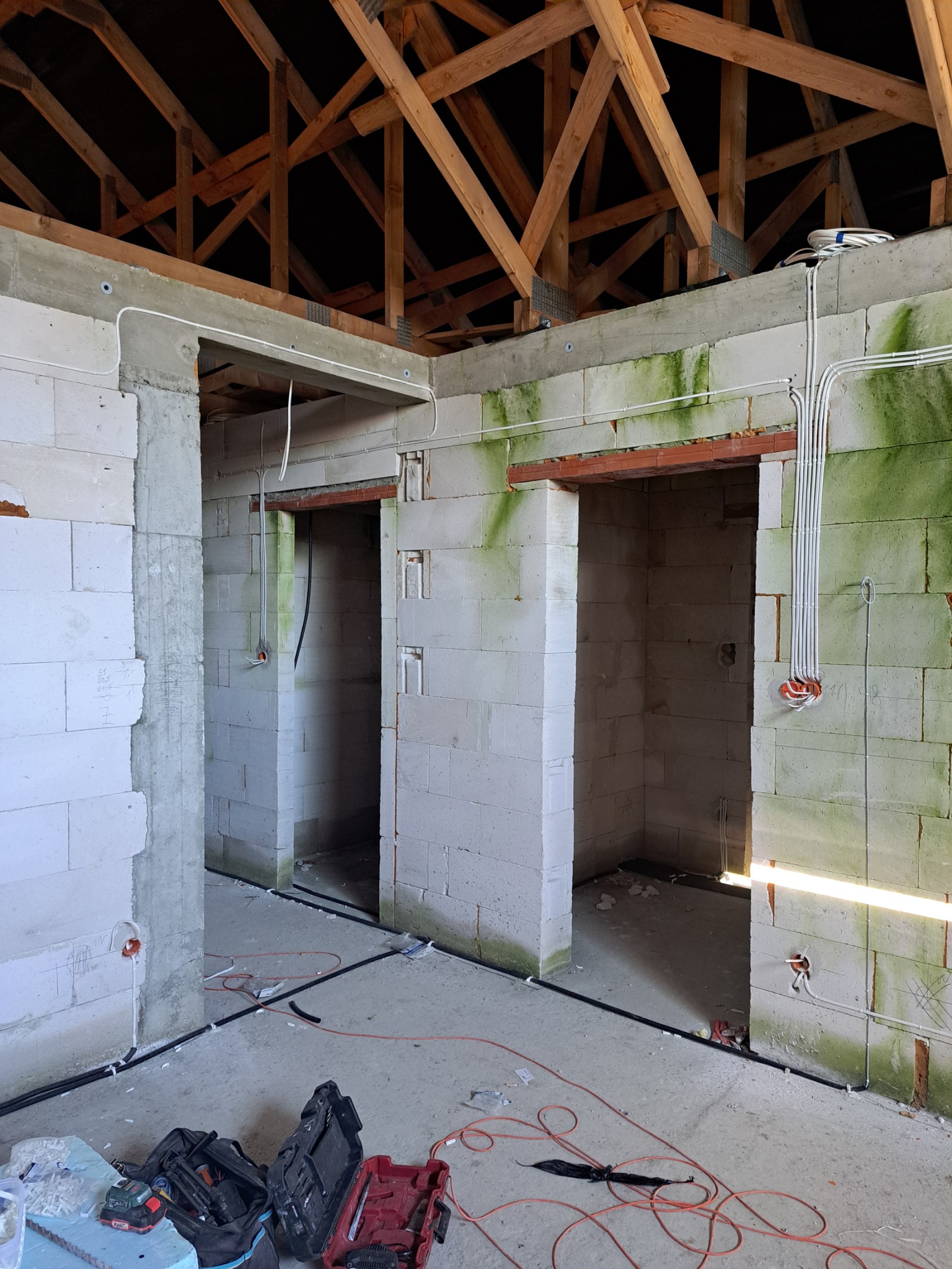 Wnętrze budynku w trakcie prac wykończeniowych, widoczne ściany z betonu komórkowego z widocznymi przewodami elektrycznymi, drewniana konstrukcja dachu oraz narzędzia na podłodze.