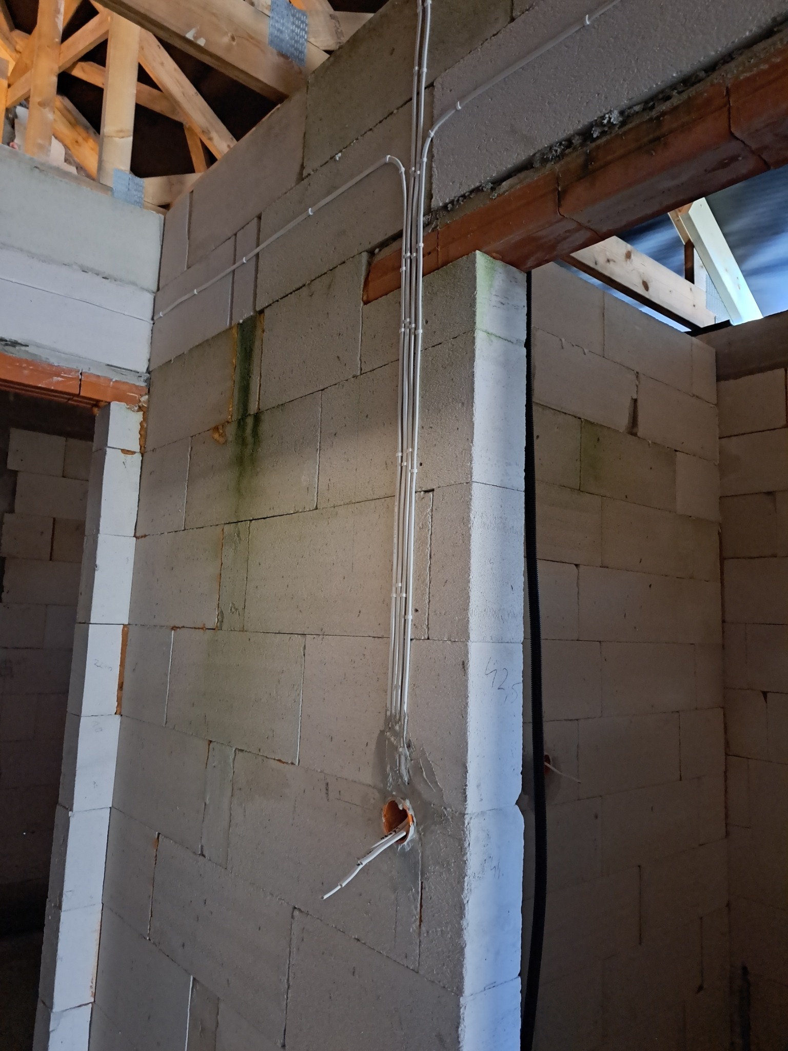 Wewnętrzna ściana z białych bloczków, z widocznymi przewodami elektrycznymi w peszlach, wychodzącymi z otworu w ścianie. Widoczne drewniane elementy konstrukcji dachu.