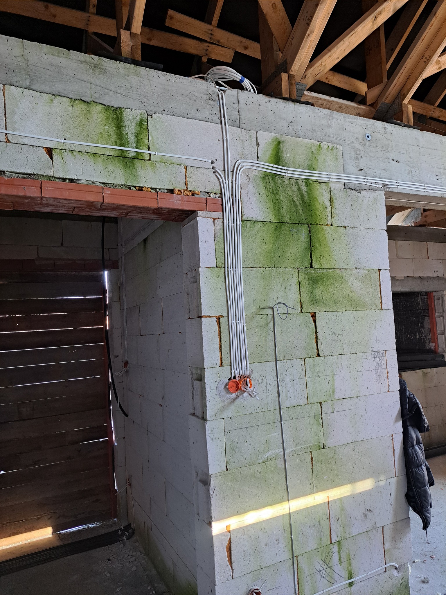 Białe przewody elektryczne ułożone pionowo na ścianie z białych bloczków z widocznymi śladami wilgoci i zielonym nalotem, pomarańczowe puszki instalacyjne widoczne u dołu, surowy stan wykończenia...