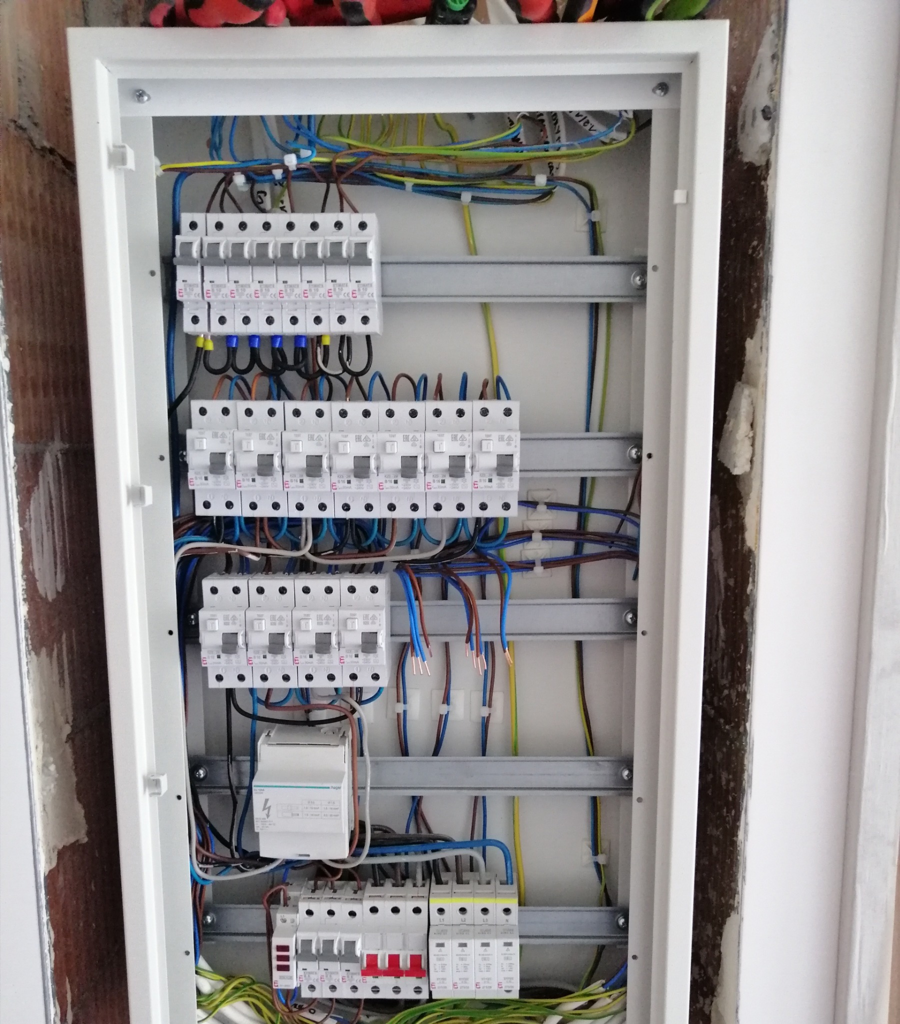 Rozdzielnica elektryczna z zamontowanymi bezpiecznikami, przewodami i innymi elementami, widoczna wnęka w ścianie z surowym wykończeniem.