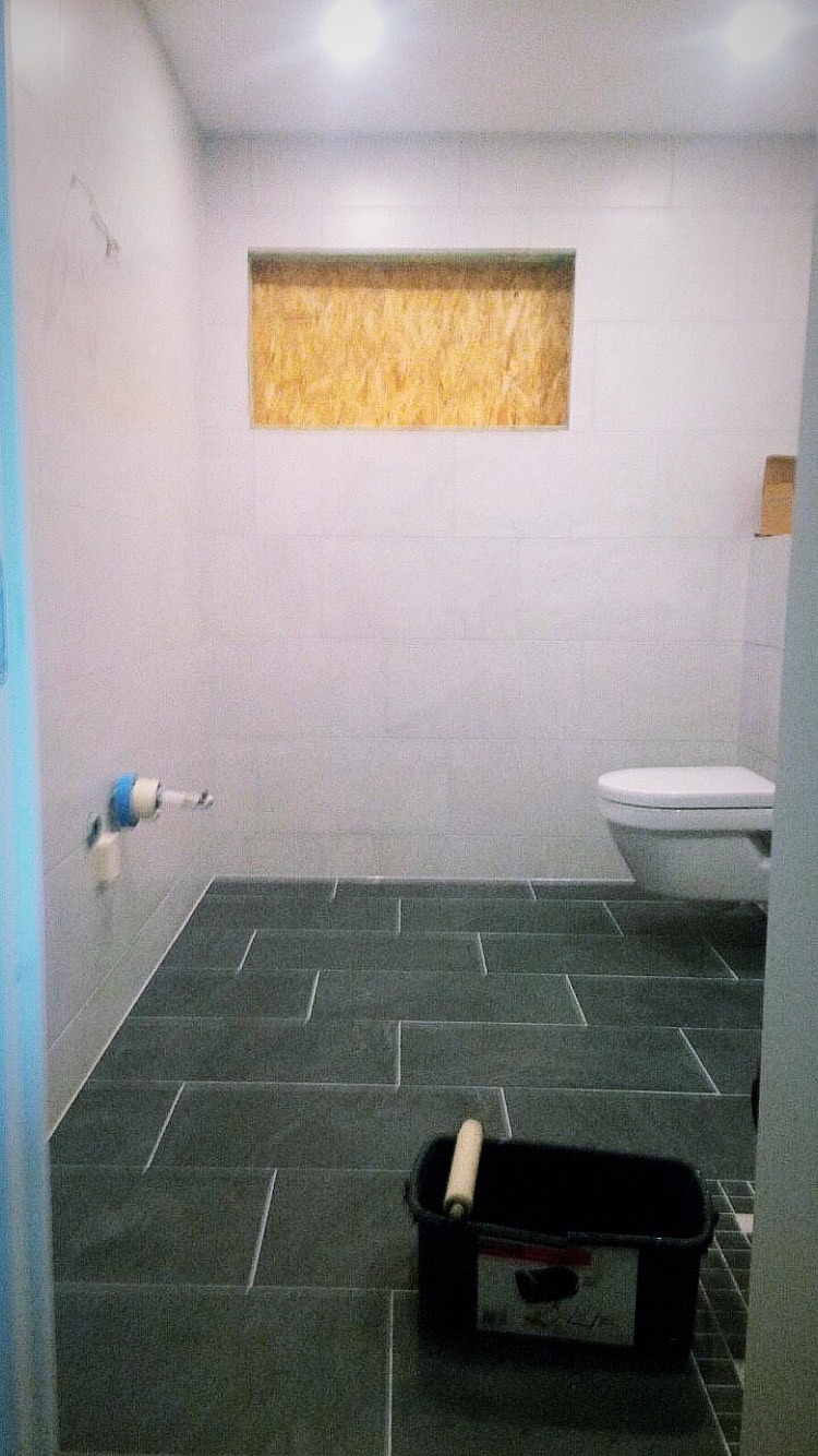 Wnętrze łazienki w trakcie prac wykończeniowych, widoczne szare płytki na podłodze, częściowo wykończone ściany, przygotowane miejsce na zabudowę, wiadro z wałkiem malarskim.