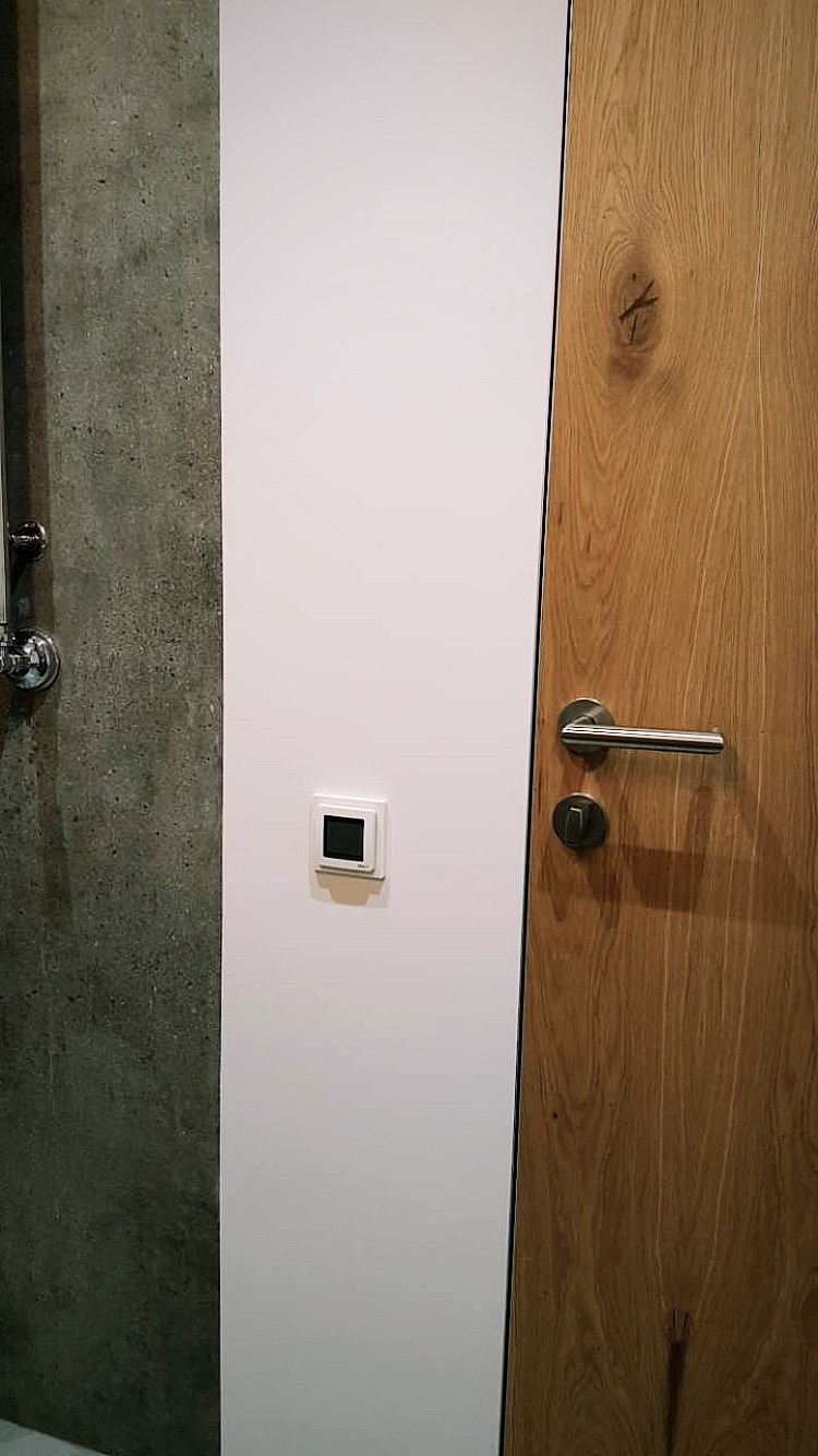 Fragment wnętrza z betonową ścianą, gładką, białą ścianą z włącznikiem dotykowym i drewnianymi drzwiami z metalową klamką.