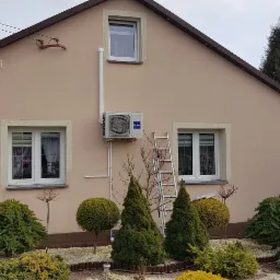 Zamontowany klimatyzator Haier na elewacji domu jednorodzinnego, widoczna drabina oparta o ścianę, zadbany ogród z ozdobnymi krzewami i figurką Świętego Mikołaja.