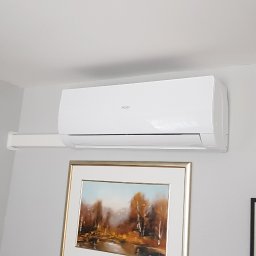 Klimathic - Jerzy Adamowicz - Biały klimatyzator Haier zamontowany na ścianie w pokoju, pod nim obraz w złotej ramie z motywem jesiennego krajobrazu.