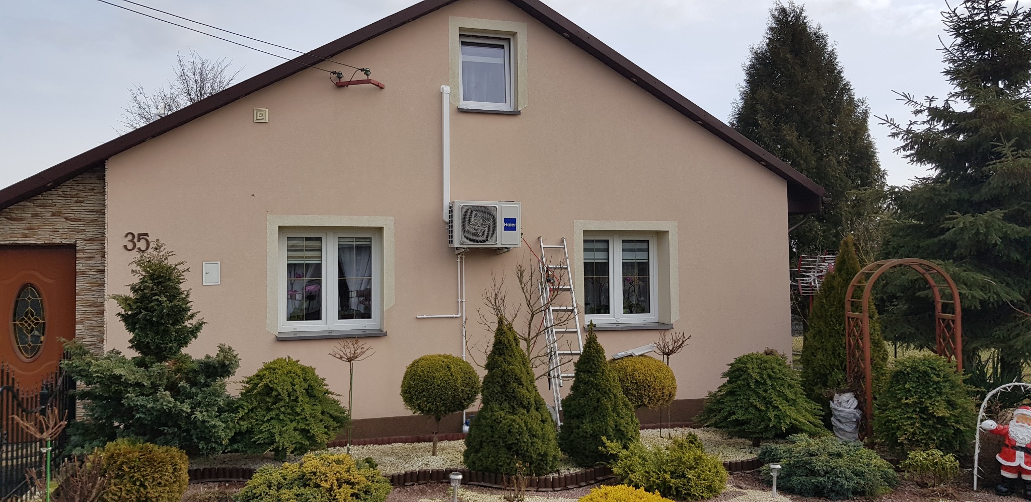 Zamontowany klimatyzator Haier na elewacji domu jednorodzinnego, widoczna drabina oparta o ścianę, zadbany ogród z ozdobnymi krzewami i figurką Świętego Mikołaja.