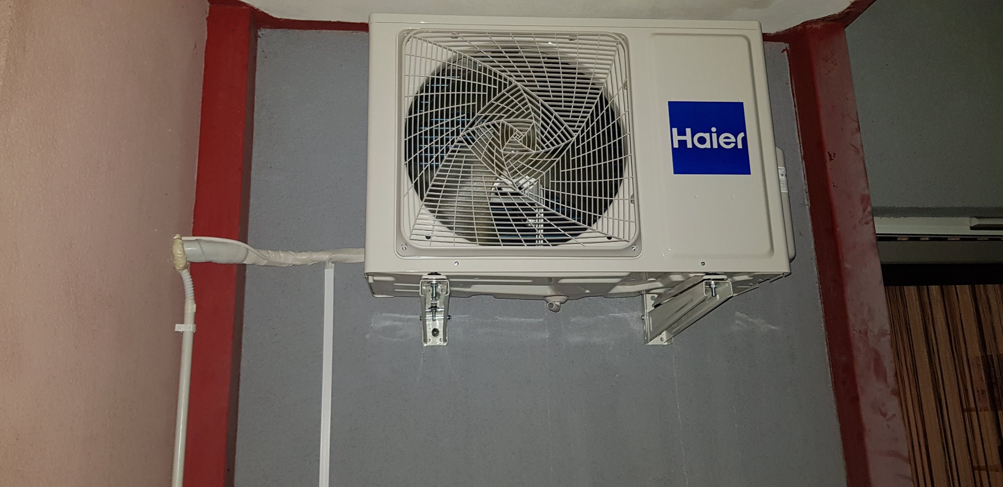 Zamontowana klimatyzacja Haier na szarej ścianie, widoczne rury odprowadzające i czerwony element konstrukcyjny budynku