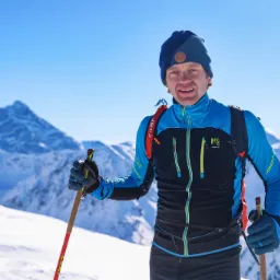 Uśmiechnięty mężczyzna w górach, ubrany w sportowy strój i czapkę, trzymający kijki narciarskie, z ośnieżonymi szczytami w tle i błękitnym niebem.