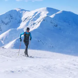 Skialpinista w niebieskiej kurtce i czarnych spodniach na tle ośnieżonych gór podczas słonecznego dnia.