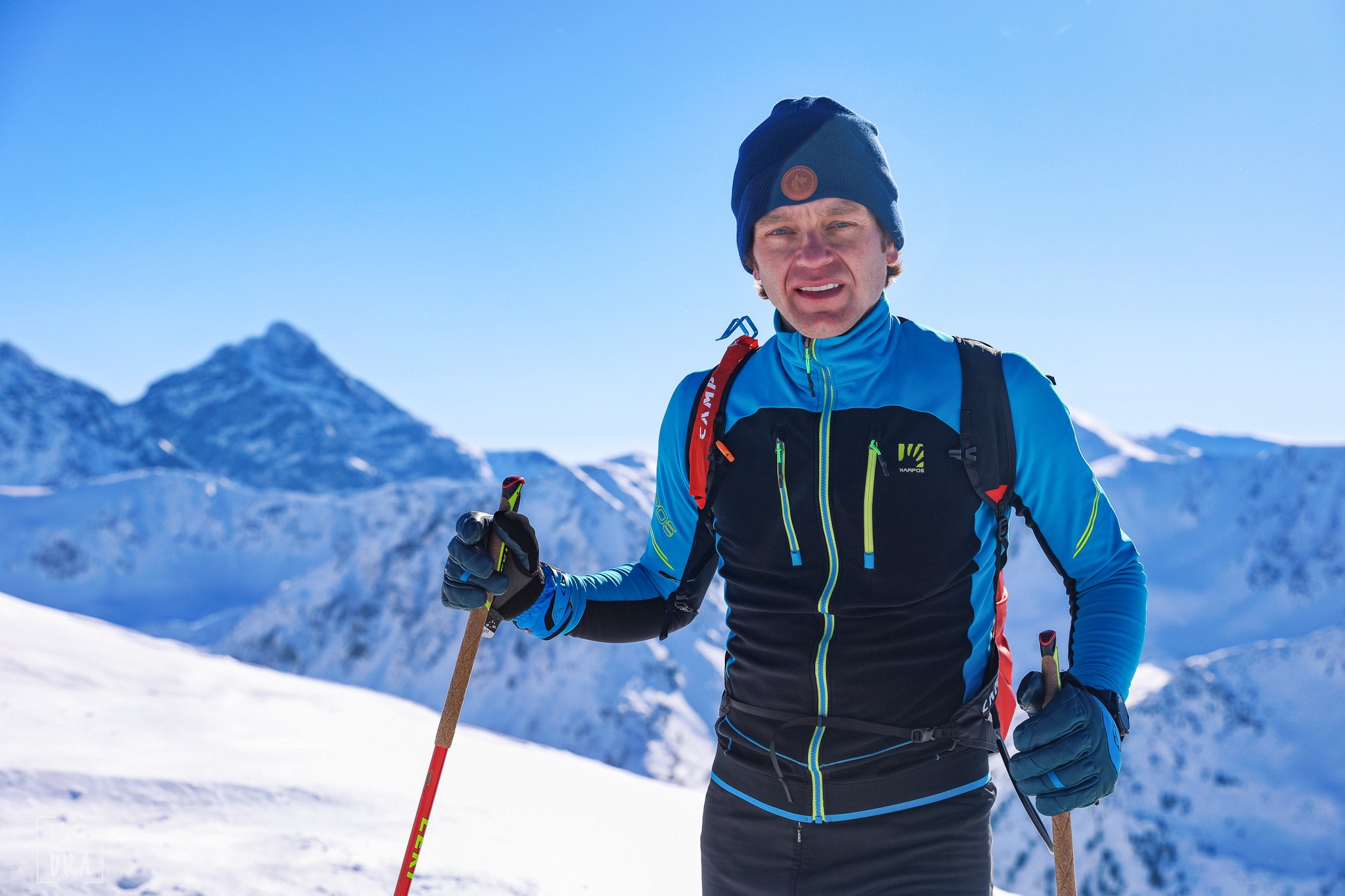 Uśmiechnięty mężczyzna w górach, ubrany w sportowy strój i czapkę, trzymający kijki narciarskie, z ośnieżonymi szczytami w tle i błękitnym niebem.