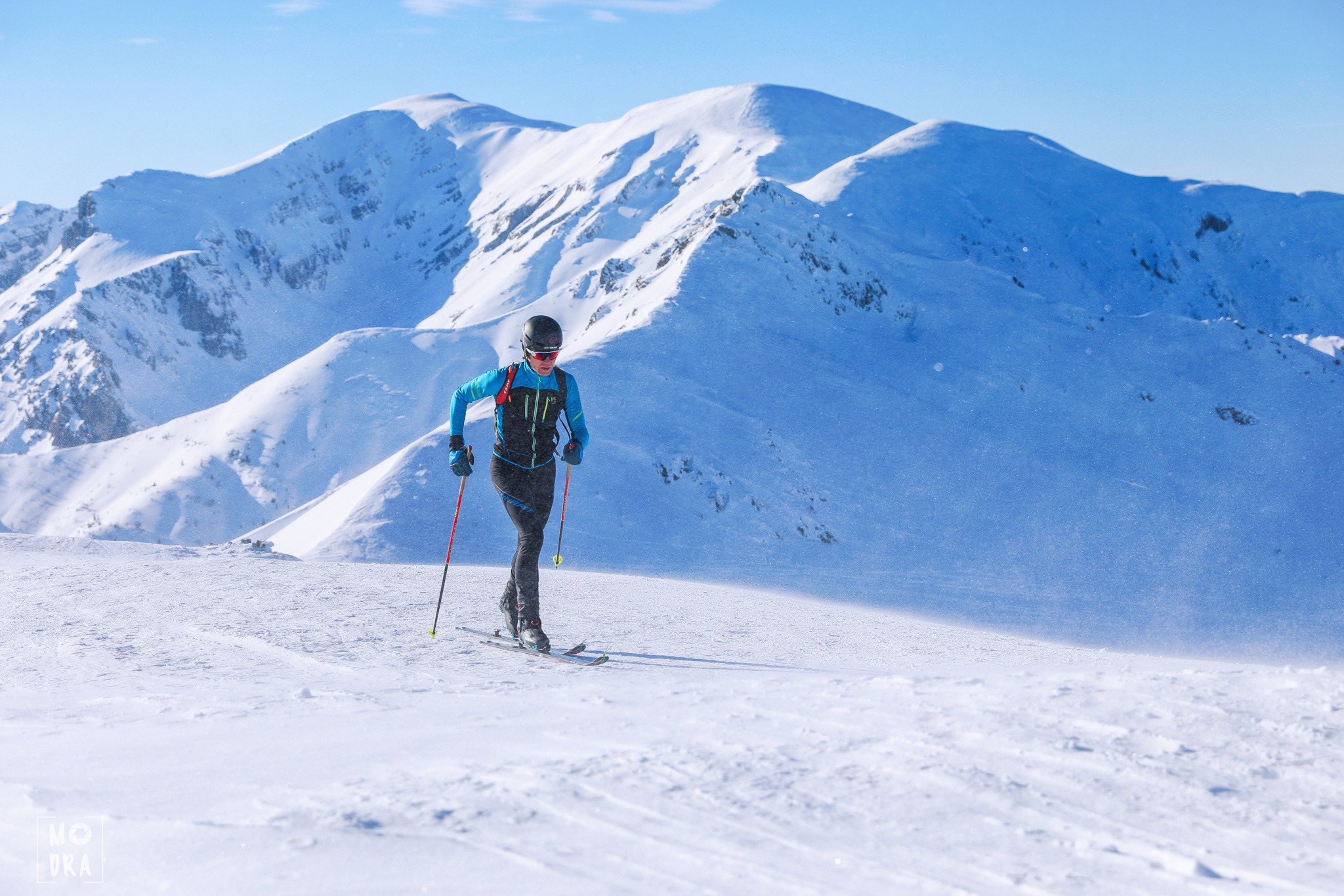 Skialpinista w niebieskiej kurtce i czarnych spodniach na tle ośnieżonych gór podczas słonecznego dnia.